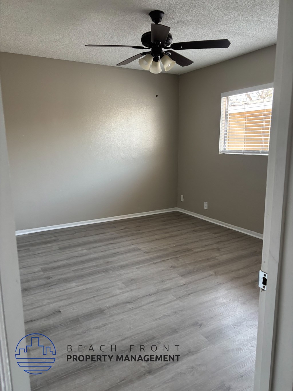 A room with a ceiling fan and light fixture, wooden flooring, and a window with blinds.